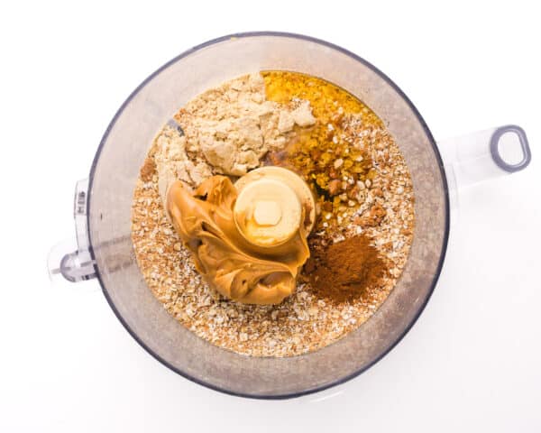 Looking down on a food processor with ingredients, such as oats, cinnamon, syrup sweetener, and peanut butter.