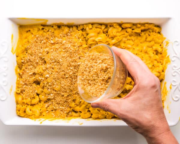 Breadcrumbs are being poured from a small bowl over Mac and cheese in a casserole dish.