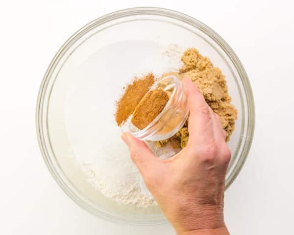 A hand pours a bowl of spices into a mixing bowl with four, sugar, and other ingredients.