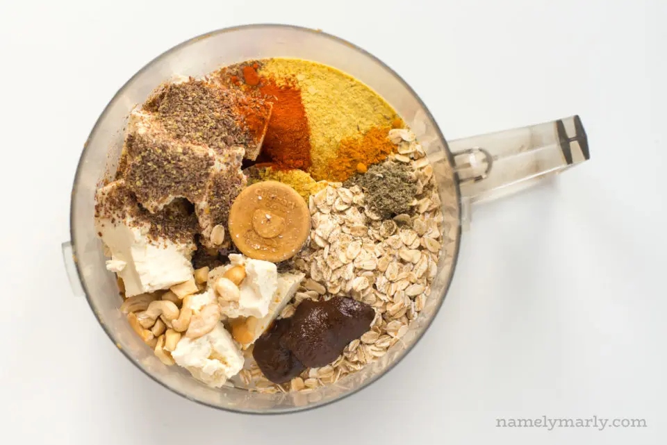 Looking down at a bowl of a food processor with ingredients in it, including oatmeal, tofu, spices, and more.