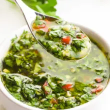 A hand holds a spoonful of chimichurri sauce, hovering over a bowl full of it. There are fresh green herbs in the background.