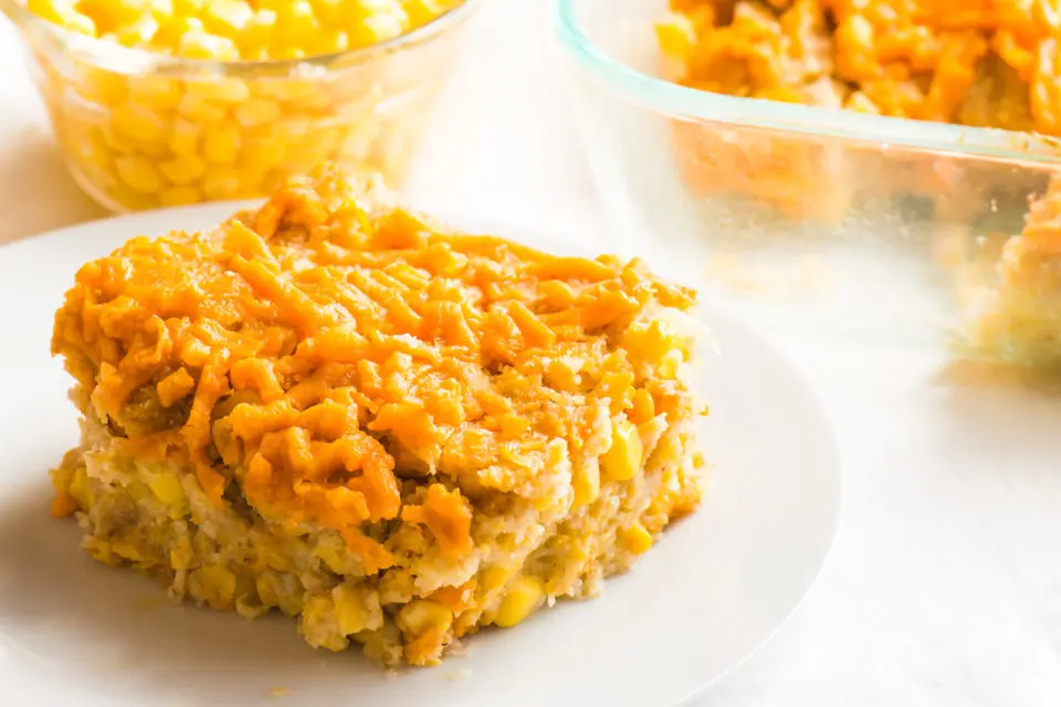 A slice of vegan corn casserole sits on a plate with the rest of the casserole in a dish behind it and corn in a bowl
