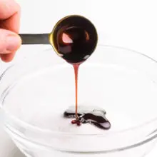 A hand holds a measuring spoon, drizzling molasses over a bowl of sugar.