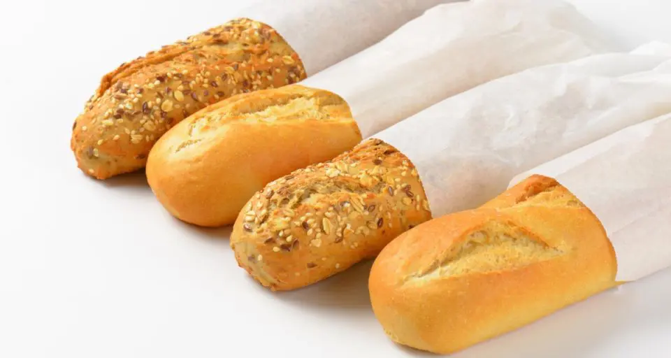 Four baguettes are rolled in white paper, with the ends of the baguettes coming out. They are sitting on a white table. Some have seeds on top, indicating they are probably whole grain.