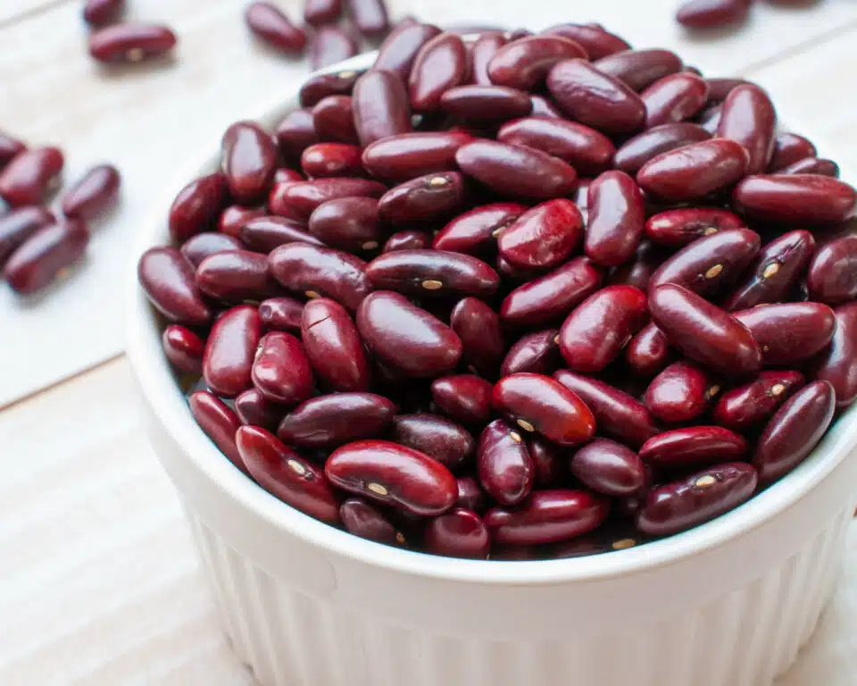 A white bowl holds shiny red kidney beans. Some beans have spilled out onto the table beside the bowl.