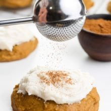Spices are being sifted over a soft pumpkin cookie with frosting on top. There's a bowl of spices and more cookies behind it.