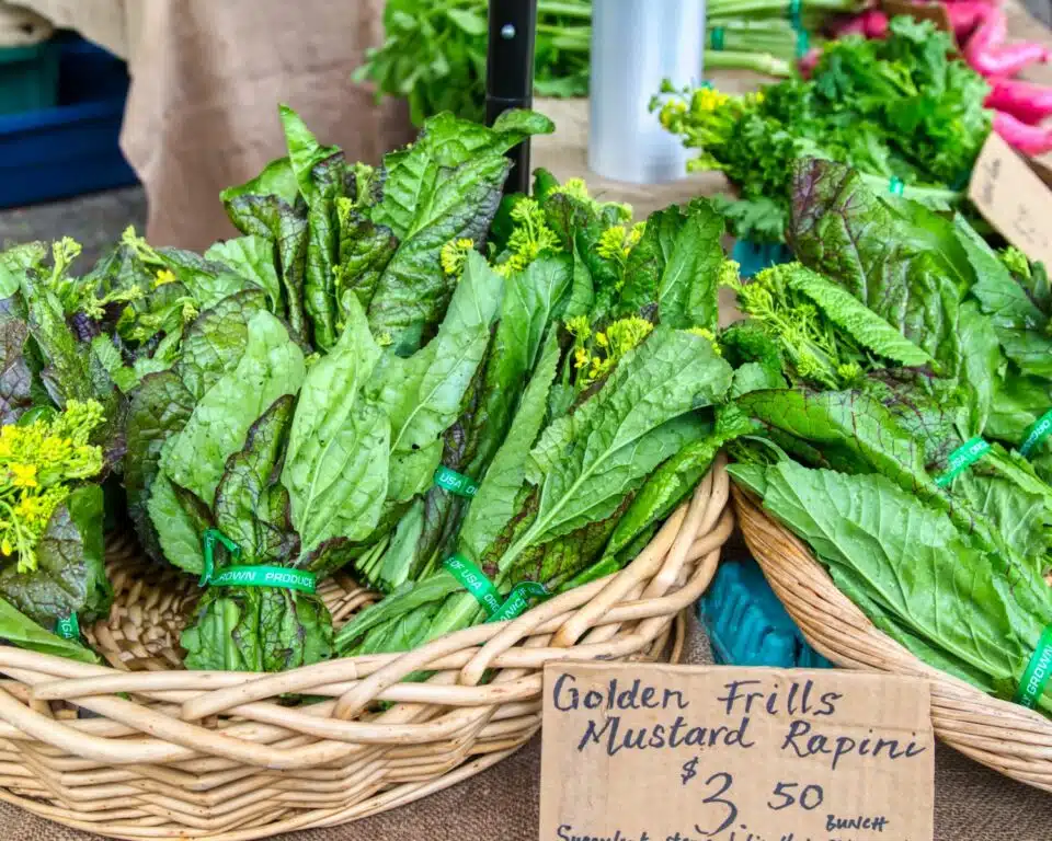 Mustard rapini bunches are in baskets and a sign sits in front, implying this image is taken at a farmer's market. The sign reads, Golden frills mustard rapini $3.50 per bunch.