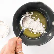 A hand pours flour over melted vegan butter in a sauce pan.