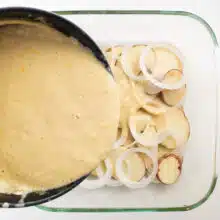 A pan of cream sauce is being poured over layers of potatoes and onions in a baking dish.