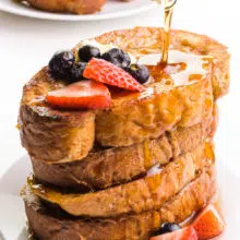 Syrup is being poured over a stack of vegan French toast. There's a plate with more French toast in the background.