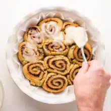 A hand holds a spoon, adding icing to freshly baked cinnamon rolls.