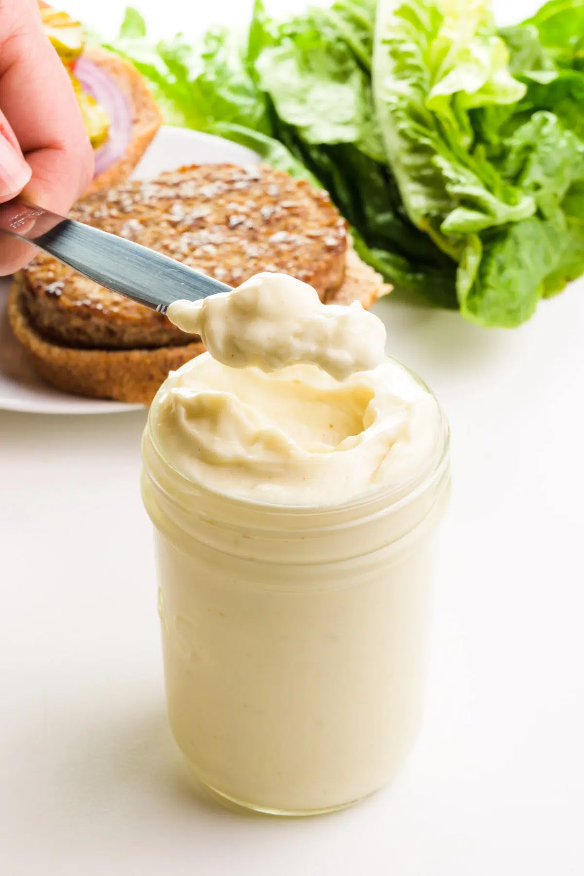 A hand holds a scoop of mayo on a knife, hovering over a jar full of it. There is a veggie burger and lettuce in the background.