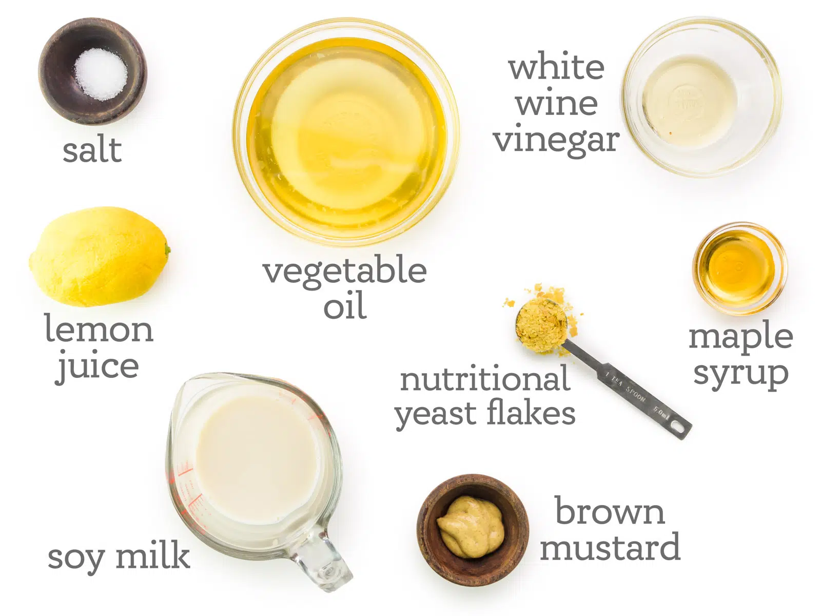Ingredients are laid out on a table. The following text is listed next to the ingredients, white wine vinegar, maple syrup, nutritional yeast flakes, brown mustard, soy milk, lemon juice, salt, and vegetable oil.