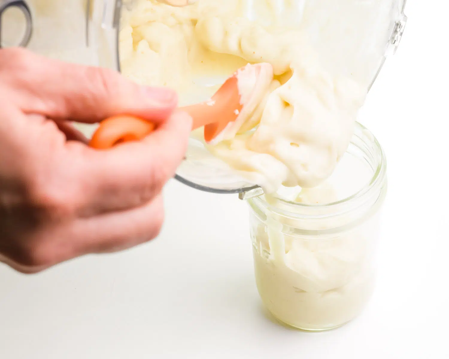 Vegan mayonnaise is being transferred from a food processor to a mason jar.
