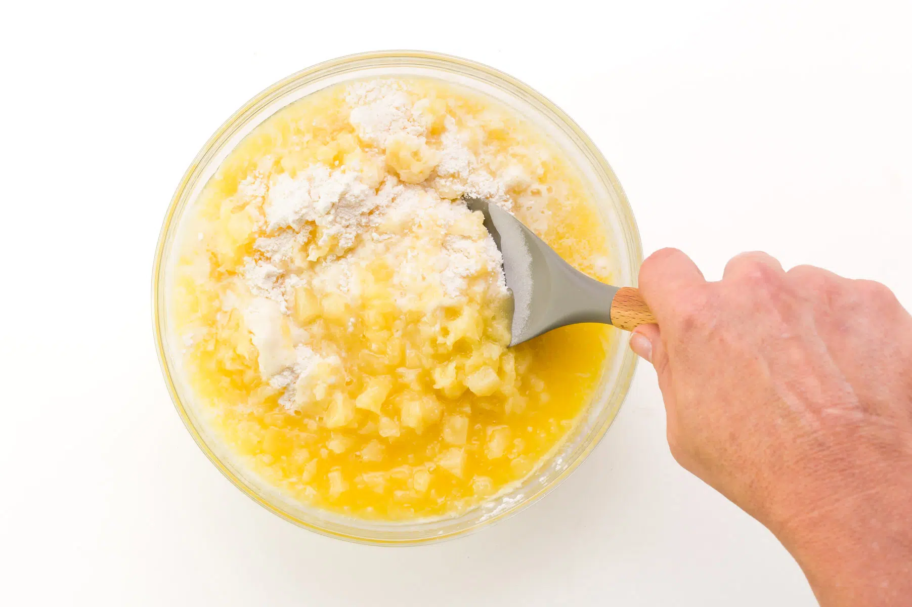A hand holds a spatula, using it to stir together cake batter with crushed pineapple.