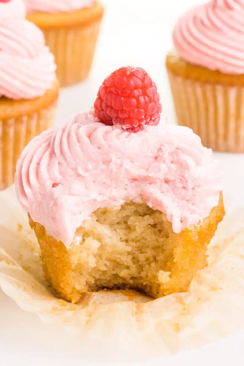 A vanilla cupcake has a bite taken out. It has a mound of pink frosting and a fresh raspberry on top. There are other cupcakes in the background.