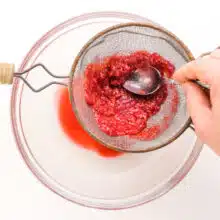 A hand pushes mashed raspberries through a fine mesh strainer into a bowl.