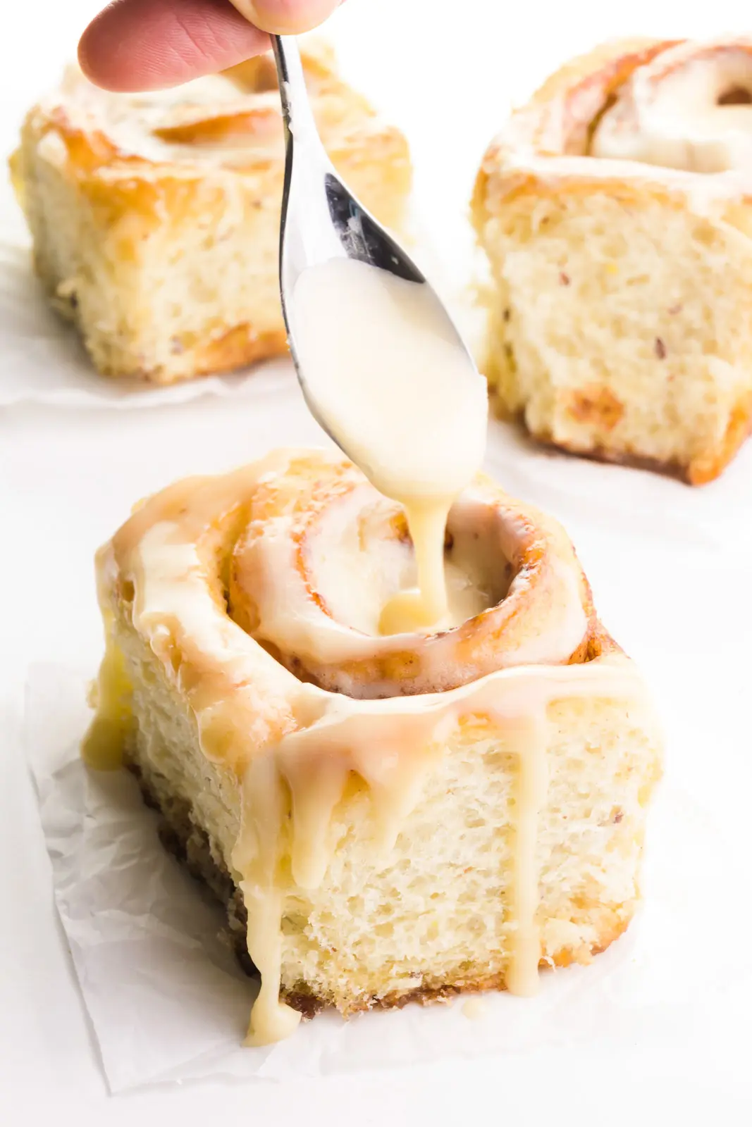 Three vegan cinnamon rolls are on a counter. A hand holds a spoon drizzling frosting over the top of one of them.