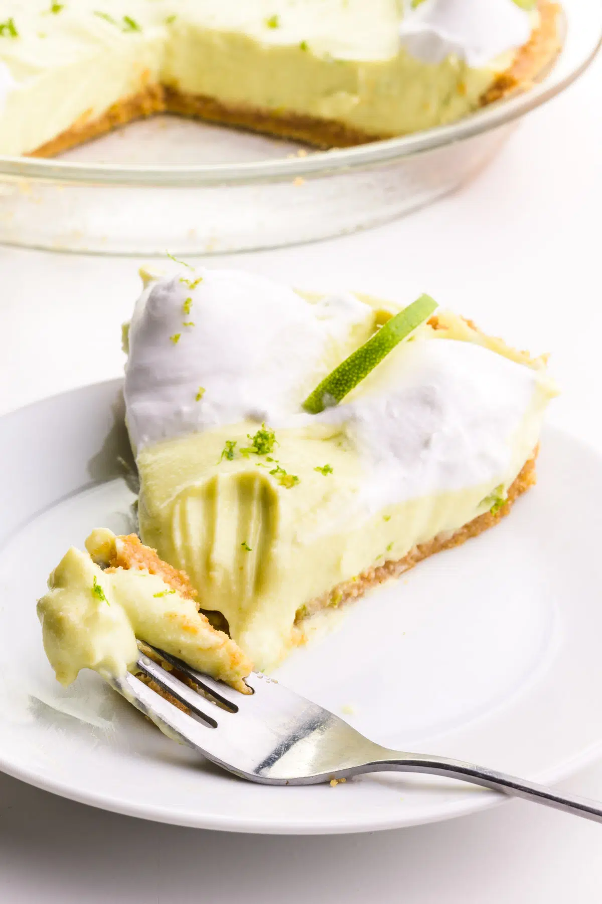 A slice of key lime pie sits on a plate with a fork in front of it with some of the pie on it. The rest of the pie is in the background.