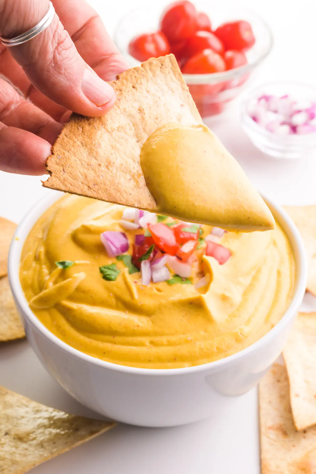 A hand holds a tortilla chip with lots of vegan nacho cheese sauce on top. The bowl of the sauce is below it and there are other ingredients like chopped tomatoes and red onions on top.